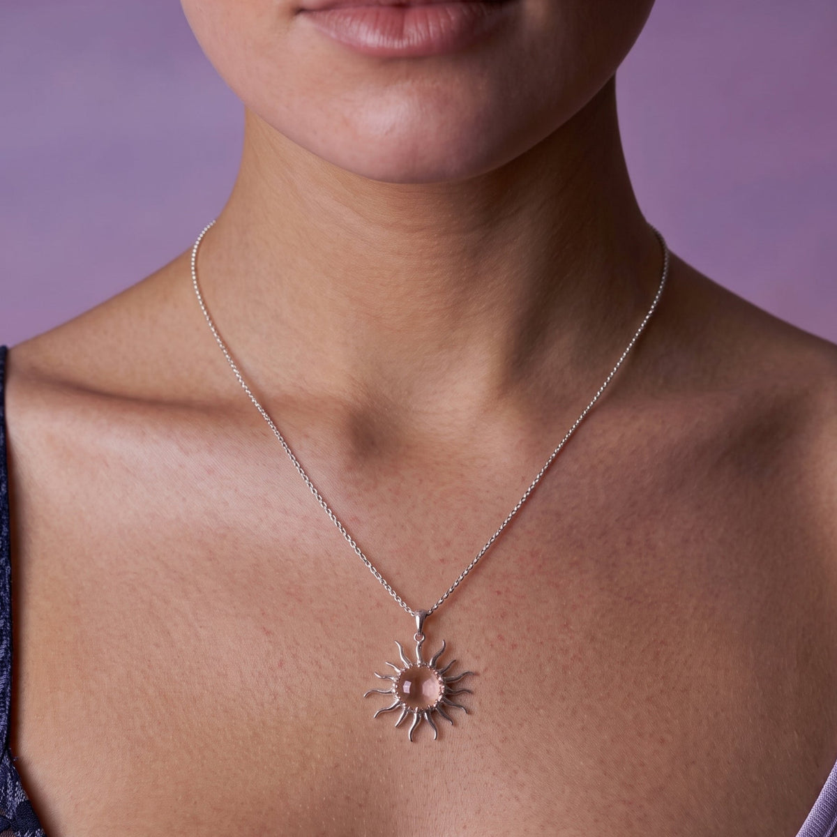 A close-up of a model wearing a delicate silver chain necklace with a sunburst pendant featuring a central pink stone, set against a soft purple background.
